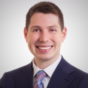 John, a white man with brown hair and blue eyes, wears a blue jacket, white shirt, and blue tie. 