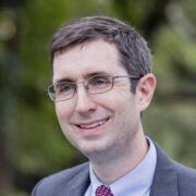 David, a white man with brown hair, wears a pale grey shirt, red tie and grey jacket. 