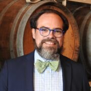 John, a white man with brown hair and a beard, wears glasses, a white shirt, black jacket and green bowtie.